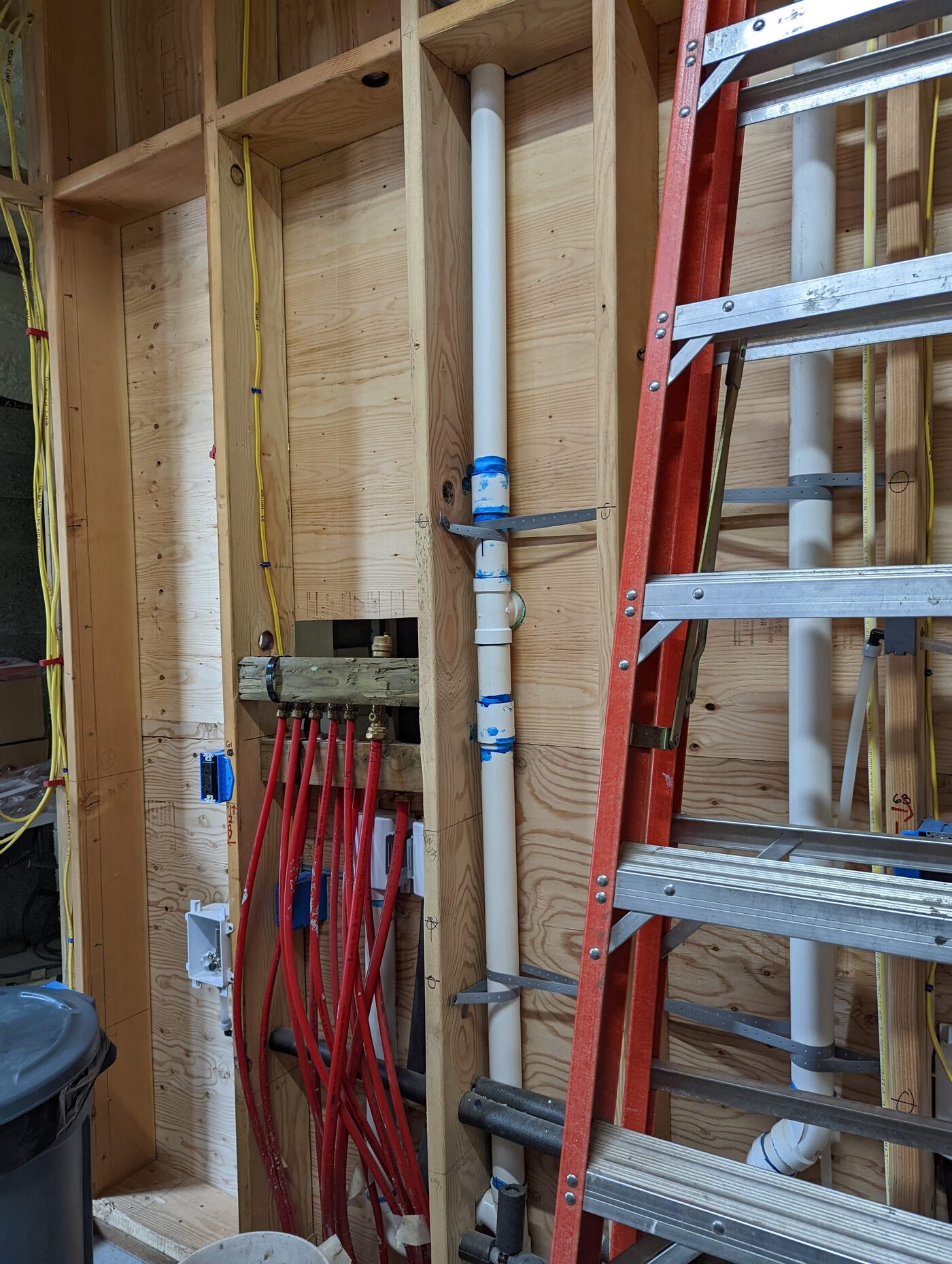 PEX manifold and drain stacks — red radiant supply lines, white drainage, yellow electrical in framed wall cavity